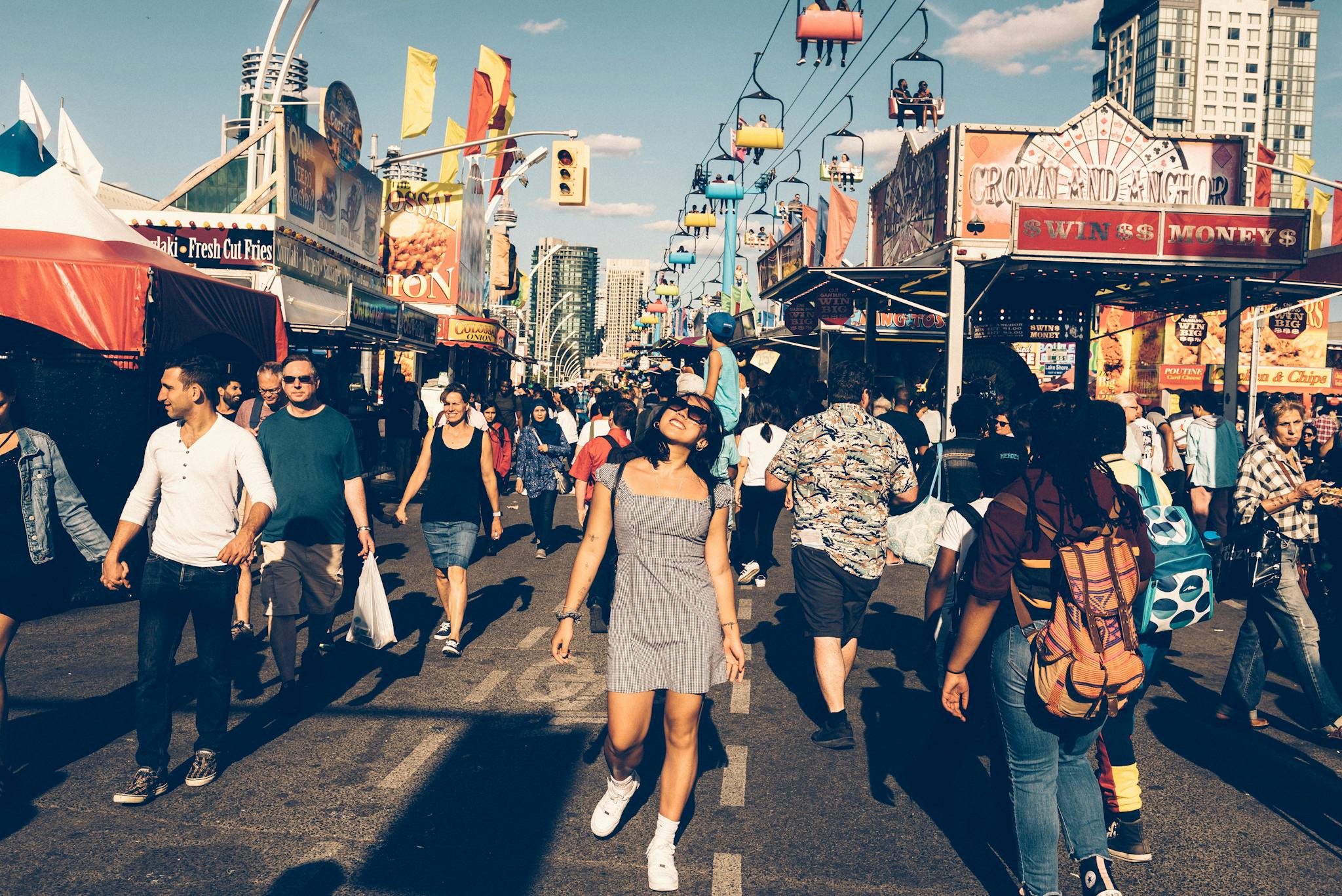 A lively street festival with diverse people enjoying a sunny day and a visible Ferris wheel.